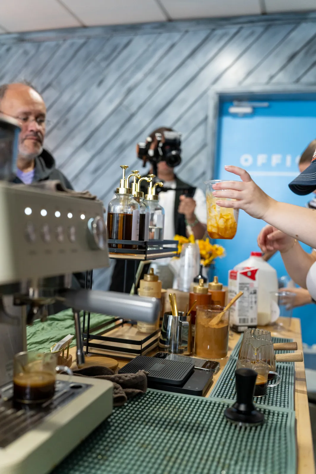 A barista hands a finished drink across the cart, the bar lined with syrups, milk and sunflowers.