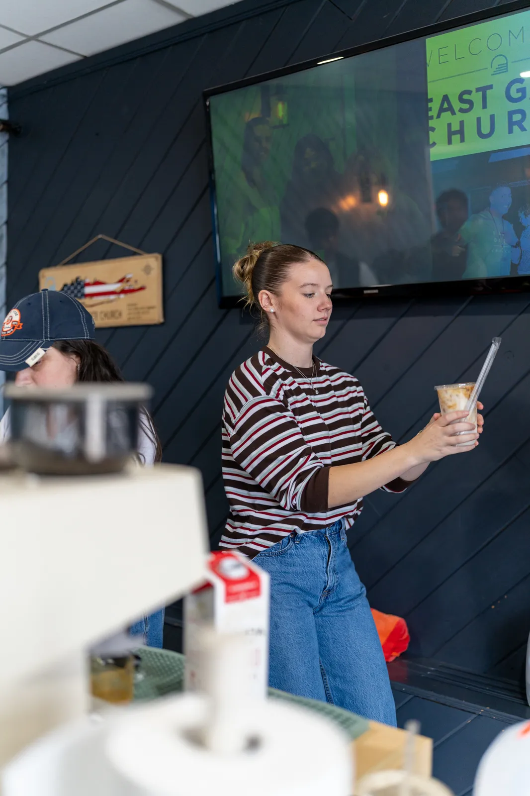A barista in a striped top holds up a finished iced drink in front of the lobby screen.