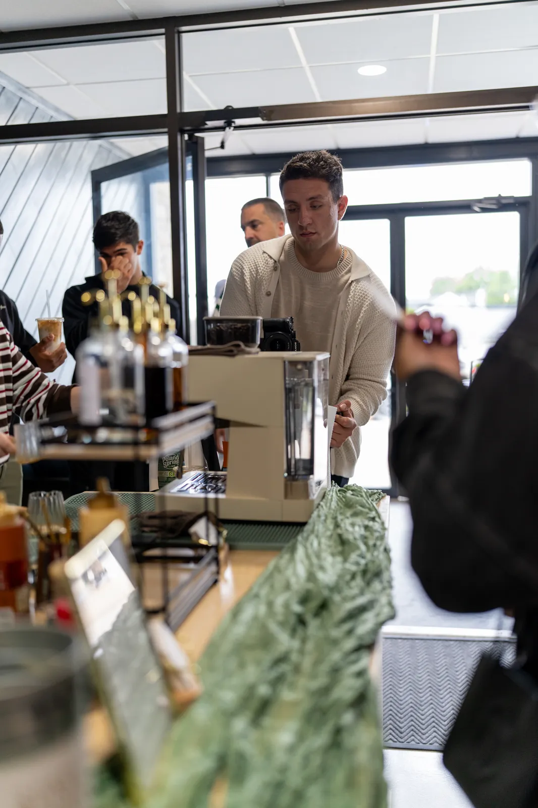A barista in a cream sweater works the espresso machine as guests wait at the cart.