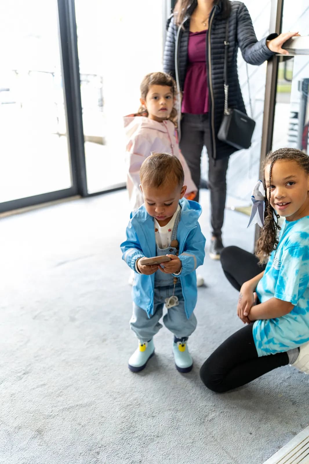 A toddler in a blue jacket walks into the lobby while an older child kneels and smiles.