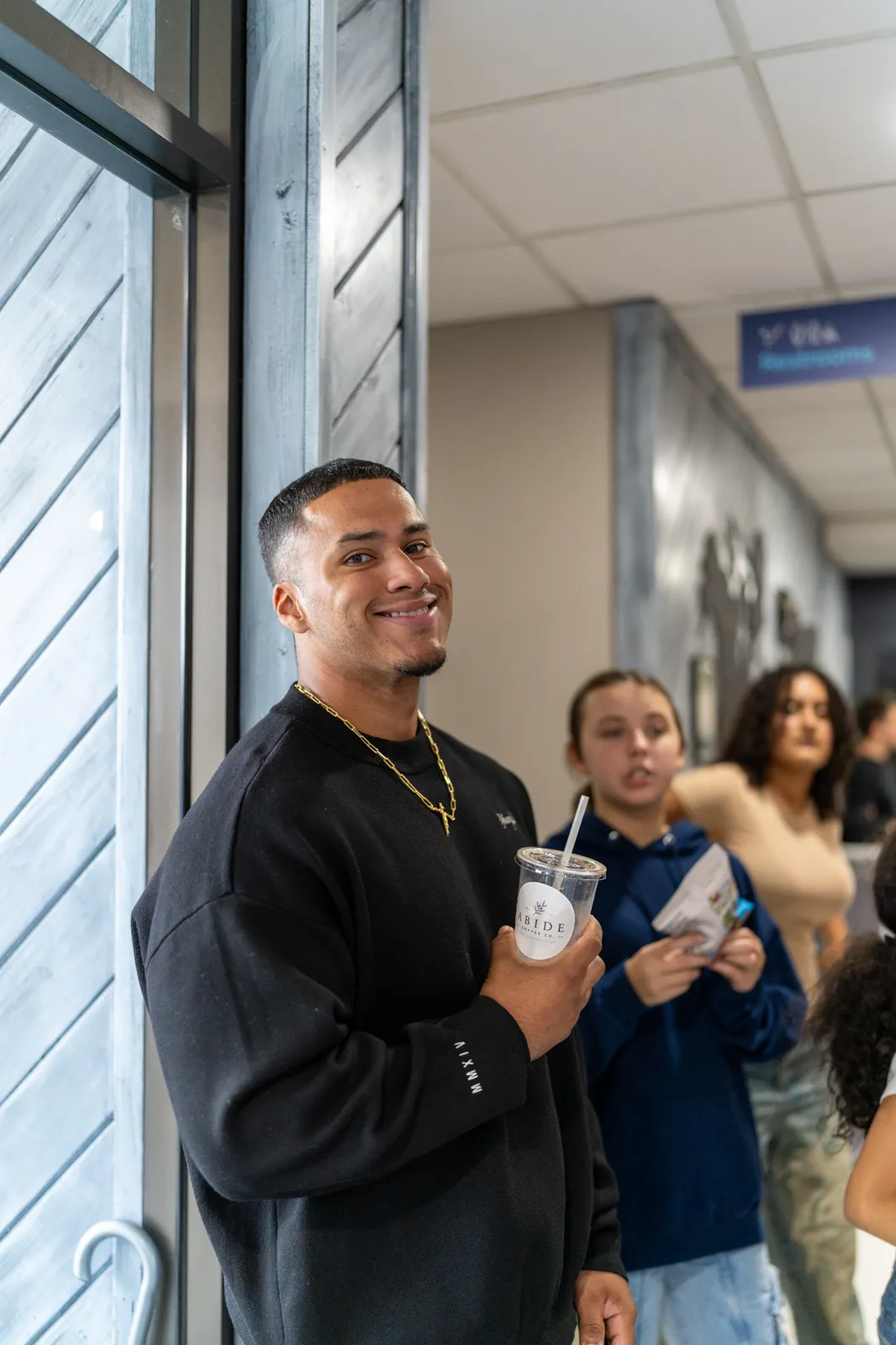 A guest in a black crewneck smiles with an iced coffee by the doorway.