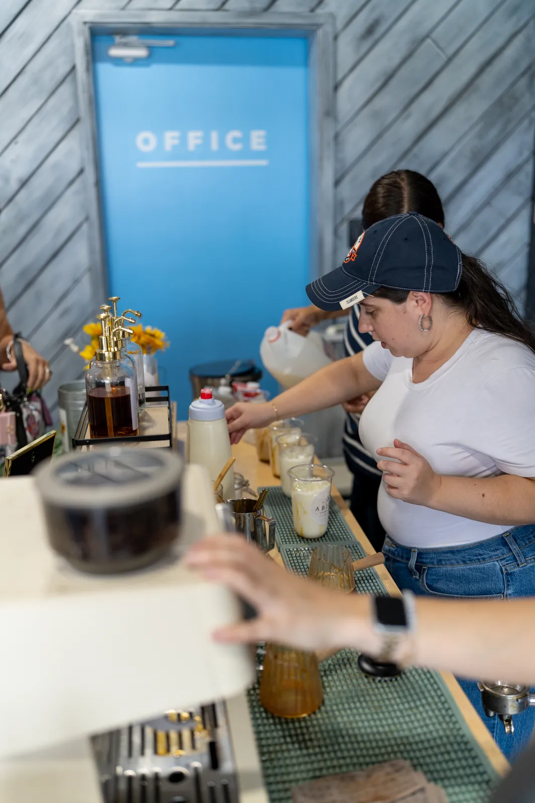 A barista in a cap pours milk at the bar beneath the blue office door.