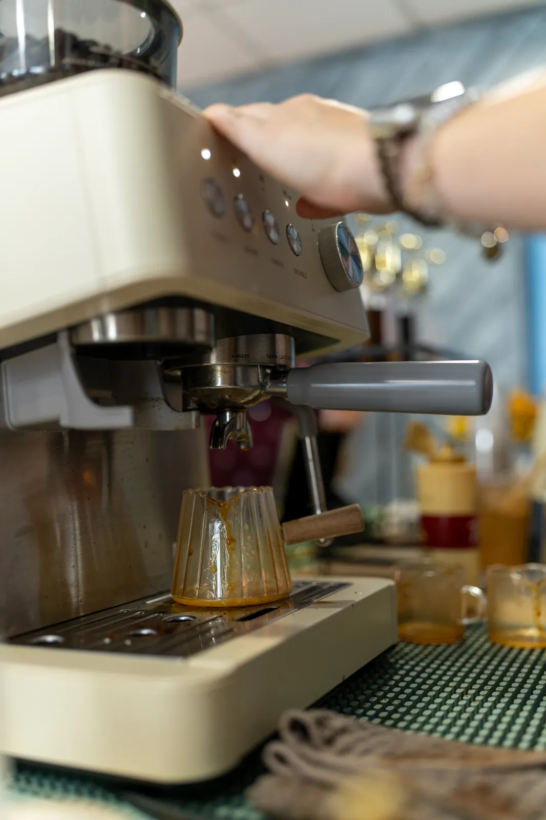 A hand rests on the cream espresso machine as a fresh shot pulls into a glass.