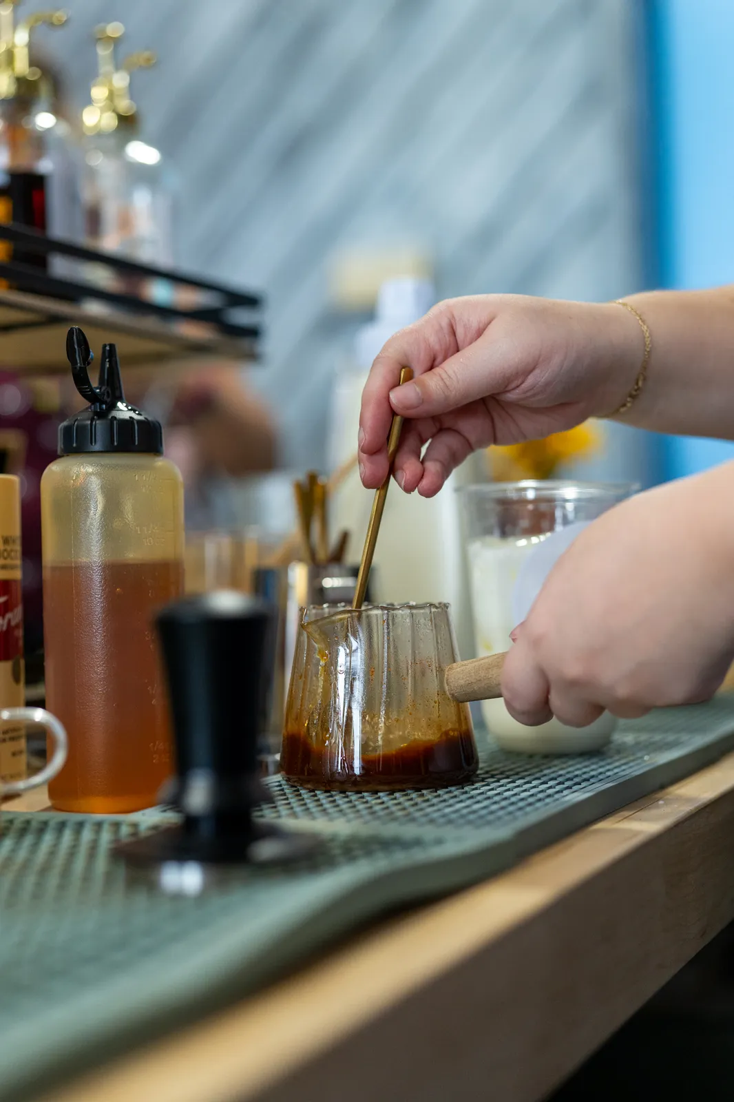 A barista stirs a drink beside a bottle of house syrup on the green bar mat.