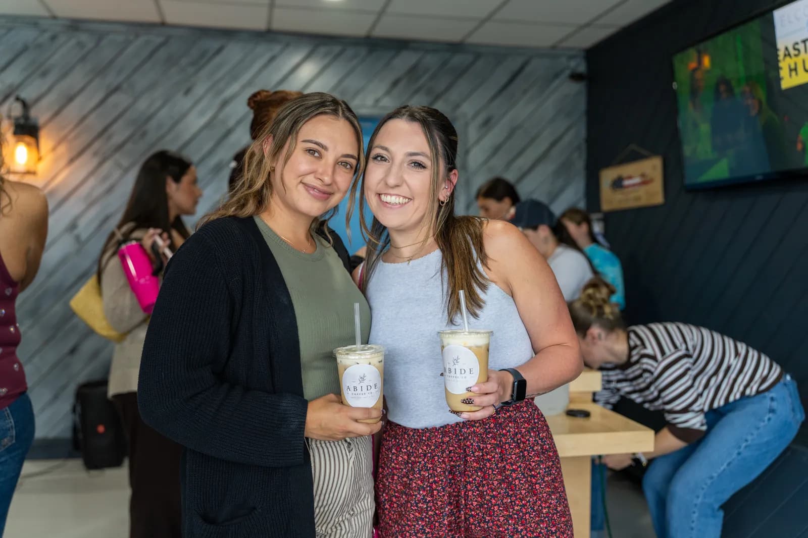 Two friends smile with their iced drinks against the shiplap wall.