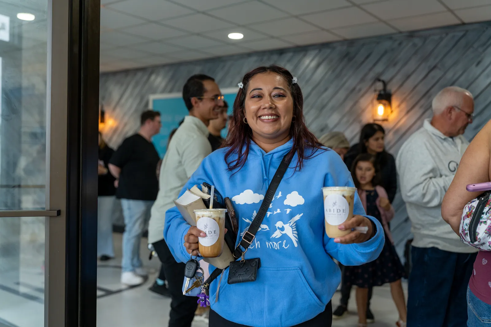 A guest in a blue hoodie beams, holding two Abide iced drinks.