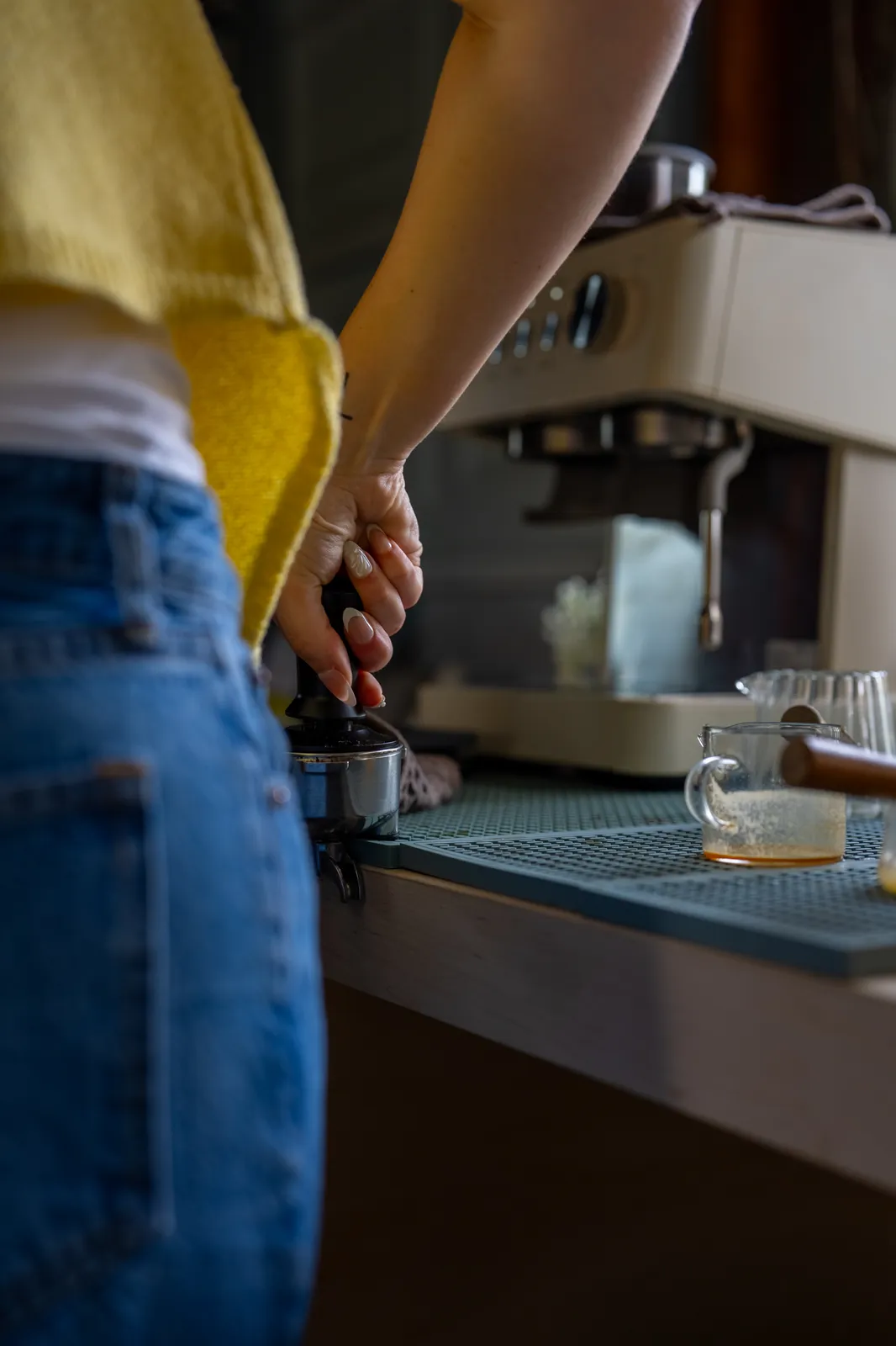 A barista in a yellow vest presses a tamper into a portafilter beside a cream espresso machine.
