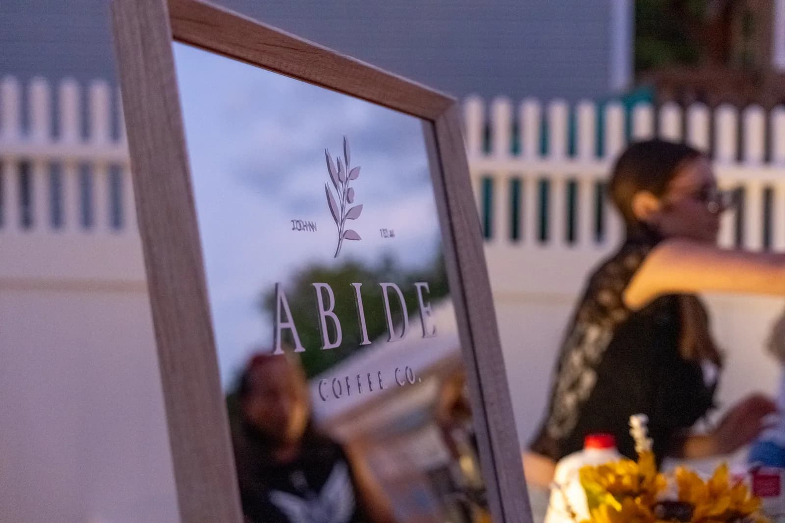 An etched Abide Coffee Co. mirror sign reflects the evening sky, with a guest in sunglasses and sunflowers at the cart behind it.