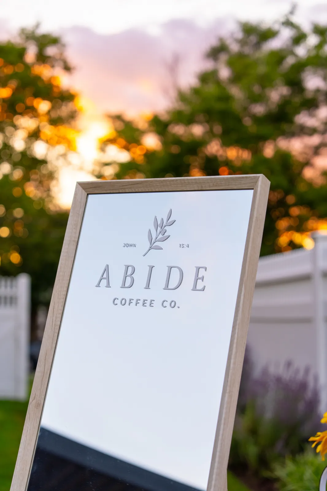 The wood-framed Abide Coffee Co. sign, marked John 15:4, stands in front of trees lit by a warm sunset.